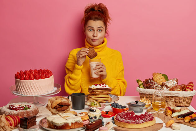 吃人 营养 热量 面包店的概念穿着黄色套头衫的姜姑娘吃美味的燕麦片饼干 喝酸奶 坐在桌上吃许多美味的蛋糕 不坚持节食甜点奶油女性