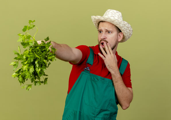 年轻年轻帅气的斯拉夫园丁穿着制服 戴着帽子 伸着香菜看着一边 手放在嘴上 隔离在橄榄绿的墙上 留着临摹的空间花园斯拉夫制服