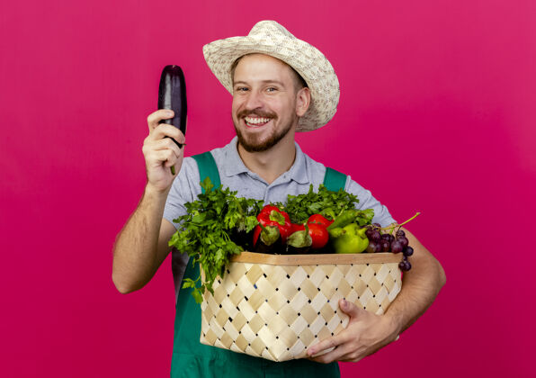蔬菜快乐的年轻英俊的斯拉夫园丁 穿着制服 戴着帽子 手里拿着一篮子蔬菜和茄子 隔离在深红色的墙上花园斯拉夫茄子