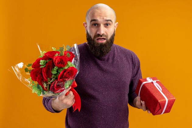 天一个留着胡子的年轻人拿着一束红玫瑰和一份礼物看着相机 站在橙色背景下惊讶地发现情人节的概念惊喜胡子概念