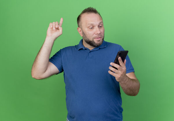 电话印象深刻的成年斯拉夫男子举着手机看着手机举起手指隔离在绿色的墙上手指斯拉夫看