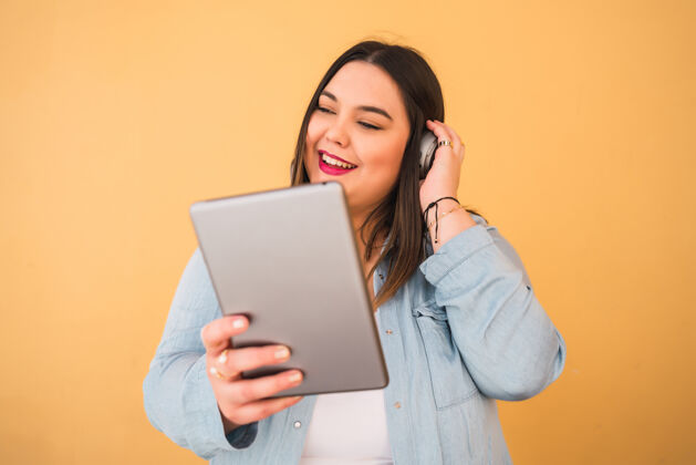 享受在黄色背景下 戴着耳机和数字平板电脑在户外听音乐的大号年轻女子的画像媒体耳机大小