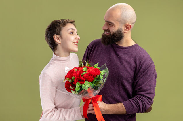 年轻一对穿着休闲服的年轻情侣站在绿色的墙边 快乐的留着胡子的男人向他微笑着庆祝情人节的女友送上一束红玫瑰花束微笑休闲