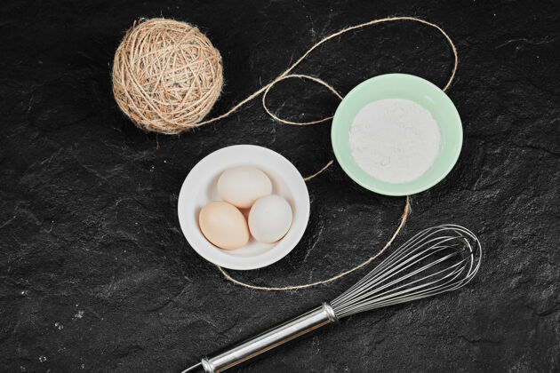 厨房瓷盘上的鸡蛋和一碗面粉放在深色的有胡须的桌子上蛋壳桌子白