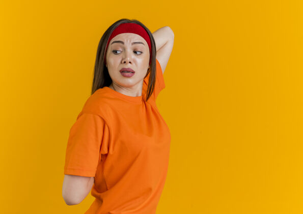 印象令人印象深刻的年轻运动女性戴着头带和腕带站在侧视图看一边保持双手放在背后练习隔离在橙色墙上复制空间站立保持侧面