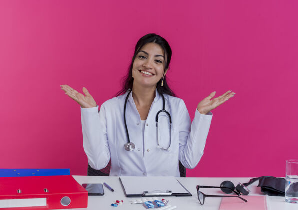 穿微笑着的年轻女医生穿着医用长袍和听诊器坐在办公桌旁 手里拿着医疗工具 两手空空的 被隔离在粉红色的墙上桌子展示空