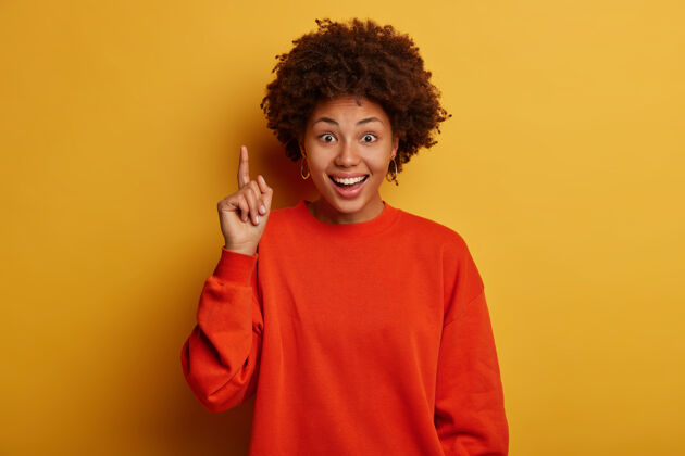 积极很高兴黑人发型的黑皮肤女人推荐很棒的产品 点起来很满意 给你看完美的宣传片 穿着红色毛衣 隔离在黄色的工作室墙上表达华丽快乐