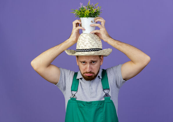 看着可怜的年轻英俊的斯拉夫园丁 穿着制服 头戴帽子 把花盆孤立在紫色的墙上头花园悲伤