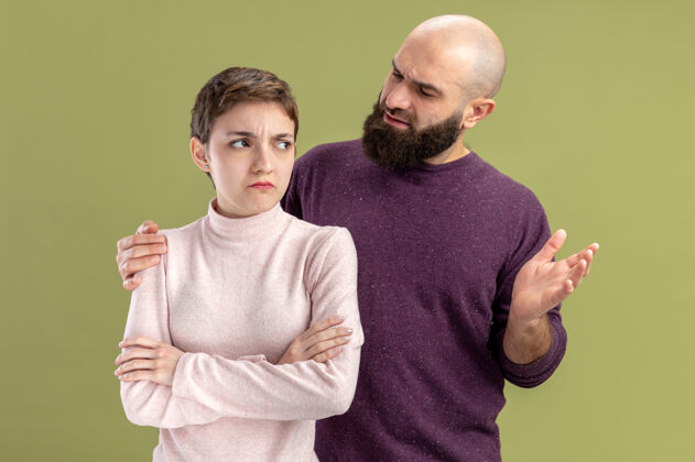 困惑一对穿着休闲服的年轻情侣 留着胡子的男人困惑地看着他严肃的 留着短发的女友 情人节的概念站在绿色的墙上胡子立场概念