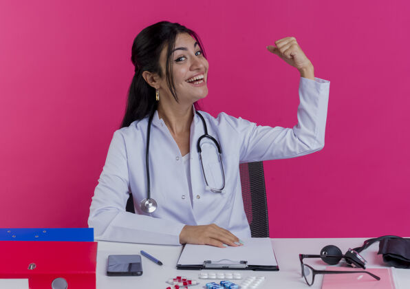 年轻快乐的年轻女医生穿着医用长袍 手持听诊器坐在办公桌旁 手放在办公桌上 做着被隔离在粉色墙上的强烈手势长袍坐着工具