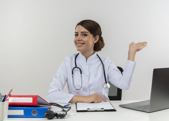 办公桌面带微笑的年轻女医生穿着医用长袍 听诊器坐在办公桌旁 手里拿着医疗工具和笔记本电脑 白墙上空空如也空长袍坐着