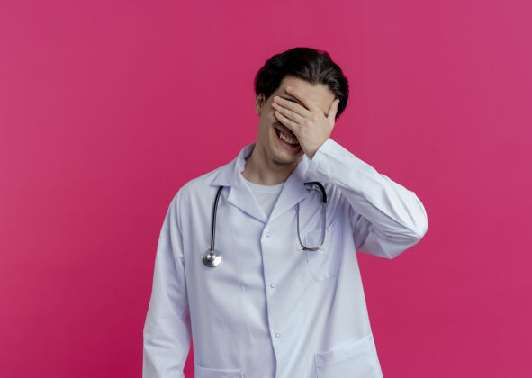 长袍面带微笑的年轻男医生 穿着医用长袍 用听诊器捂住眼睛 用手隔离在粉红色的墙上 留有复印空间微笑医生穿