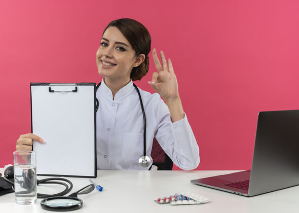 年轻面带微笑的年轻女医生穿着医用长袍 听诊器坐在办公桌旁 带着医疗工具和笔记本电脑 在粉红色的墙上贴着“剪贴板”和“一切正常”的牌子长袍穿医疗