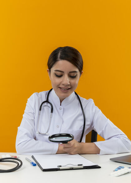 穿高兴的年轻女医生穿着医用长袍和听诊器坐在办公桌旁 拿着医疗工具和笔记本电脑 透过隔离在黄色墙上的放大镜看着剪贴板办公桌剪贴板笔记本电脑