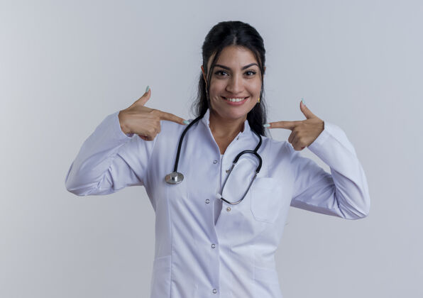 女性面带微笑的年轻女医生穿着医用长袍 用听诊器指着隔离在白墙上的自己医学医生指着