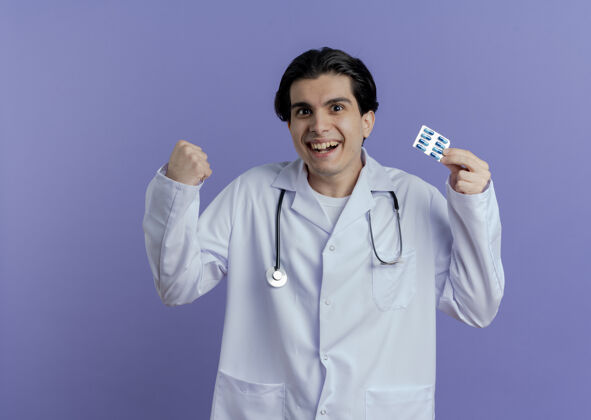 欢乐快乐的年轻男医生穿着医用长袍和听诊器 展示着一包胶囊在做“是”的手势 隔离在紫色的墙上 留有复印空间穿着男性医疗
