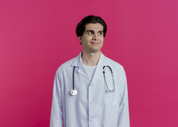 壁板高兴的年轻男医生穿着医用长袍和听诊器 看着隔离在粉红色墙上的一面有复印空间听诊器请穿长相