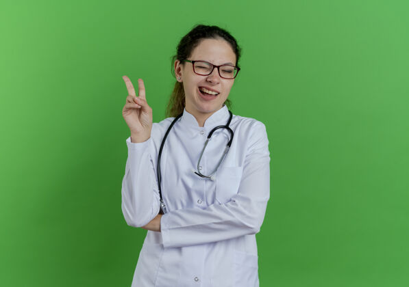 医生自信的年轻女医生穿着医用长袍 戴着听诊器 戴着眼镜 做着和平的手势 在绿色的墙上眨着眼睛 隔离着复制空间医疗眼镜穿