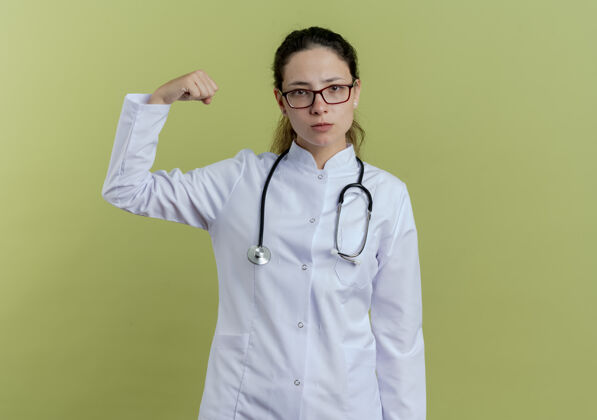 展示自信的年轻女医生穿着医用长袍 戴着听诊器和眼镜 显示出强烈的姿态隔离在橄榄绿的墙上医生长袍听诊器