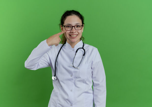 指点快乐的年轻女医生 穿着医用长袍 戴着听诊器和眼镜 手指贴在脸颊上 隔离在绿色的墙上 留着复印空间穿年轻手指
