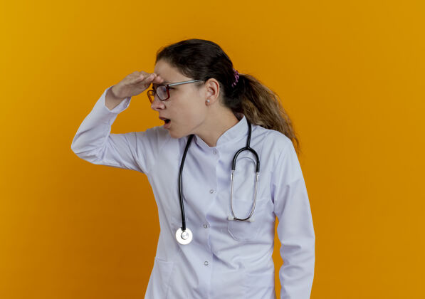 医学看着身边惊讶的年轻女医生穿着医用长袍 戴着听诊器 戴着眼镜 用手隔着橘黄色的墙遥望远方橙色穿医生