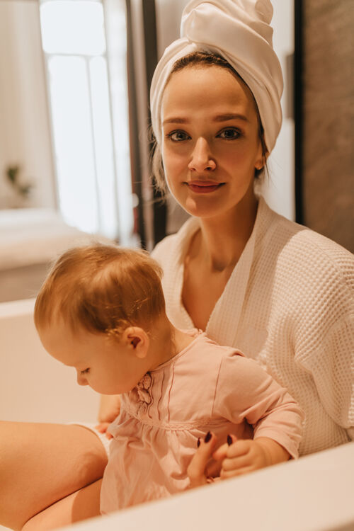 淋浴头上裹着毛巾 穿着浴衣的漂亮女孩看着镜头 坐在浴室里拥抱着她的女儿成人长袍妈妈