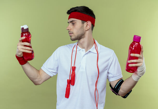 运动戴着头巾 脖子上系着跳绳 手里拿着两瓶水的年轻运动型男子 看上去很困惑 试图做出选择而不是绿色人脖子周围