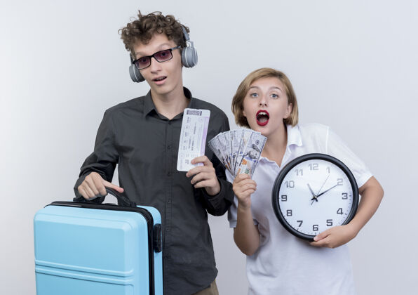 手提箱一对年轻的游客戴着耳机 拿着手提箱和机票 坐在女友旁边 手里拿着现金和挂钟 惊讶地看着白色站着情侣飞机