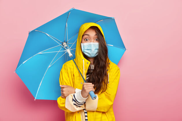 面具惊讶的黑发亚裔女子戴着医用口罩 被传染病保护着 穿着黄色雨衣 撑着雨伞脸女性兜帽