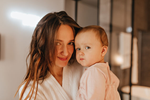 漂亮年轻微笑的妇女抱着小女儿的特写照片婴儿和母亲在公寓里合影欢呼浴袍女人