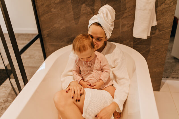 毛巾穿着浴衣和毛巾的年轻女士躺在浴室里可爱的孩子坐在妈妈身上玩耍小欢呼浴室