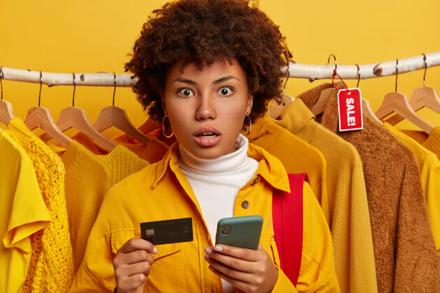 零售一位非洲发型的女士穿着黄色衬衫 在衣架上摆姿势 手里拿着现代手机和信用卡青少年电话设备