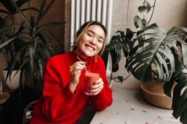 毛衣身着红色超大号毛衣的快乐女士的肖像 手里拿着一杯鲜榨西瓜汁 坐在咖啡馆靠着植物墙漂亮餐厅微笑