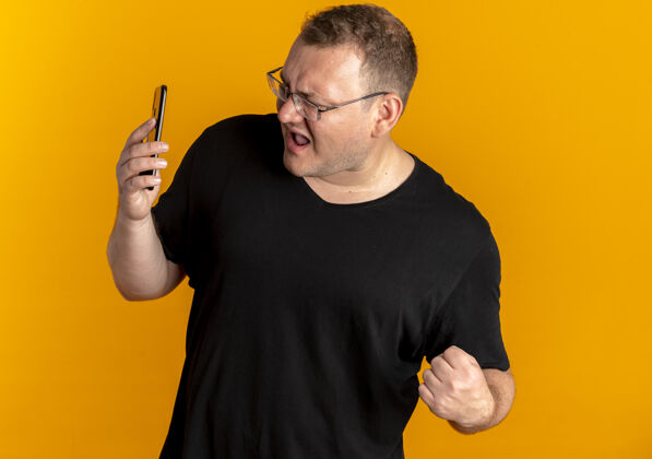手机一个戴眼镜的胖男人穿着黑色t恤看着他的智能手机屏幕握紧拳头非常难过的橙色T恤男人穿