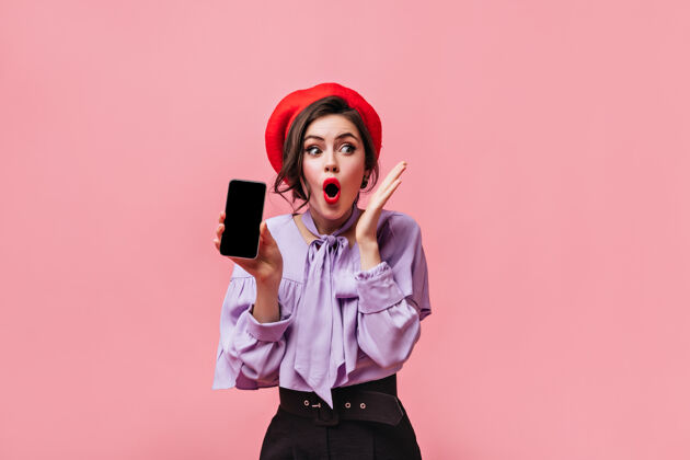年轻一位戴着红帽子 身穿淡紫色上衣的女士手持黑色智能手机 在粉色背景下摆出惊艳的姿势成人女孩衬衫