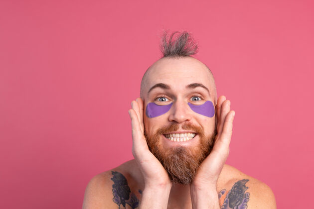 阳性欧洲帅哥留着胡子纹身上身的男人戴着紫色眼罩对着镜头摆粉红色的姿势房子化妆品凝胶