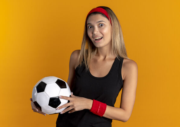 橙色身穿黑色运动服 头戴红色头巾 手持足球的年轻健身女孩惊讶地高兴地看着橙色的脸脸快乐运动装