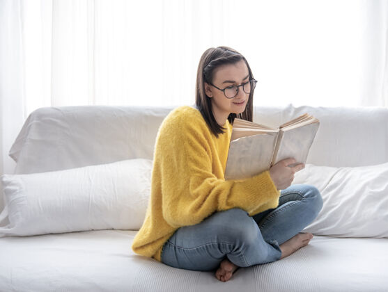 书籍穿着黄色毛衣戴着眼镜的时髦黑发女孩在家里的沙发上看书发展年轻训练