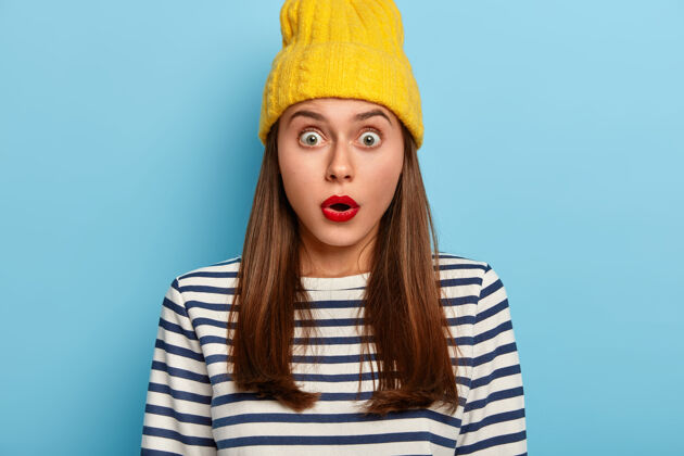 女人令人印象深刻的关注的女人对惊人的谣言作出反应 屏住呼吸 有器的眼睛 戴着黄色帽子和条纹套头衫不能相信惊讶的消息 有红唇 最小的化妆品害怕印象黑发