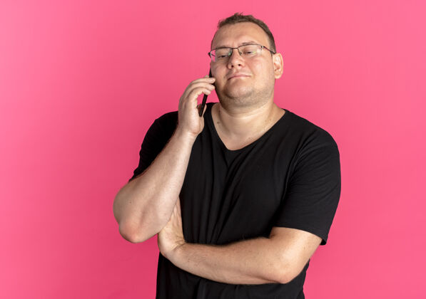 超重戴眼镜的超重男子身穿黑色t恤 一边微笑着一边用手机交谈 而不是粉色T恤男士粉色