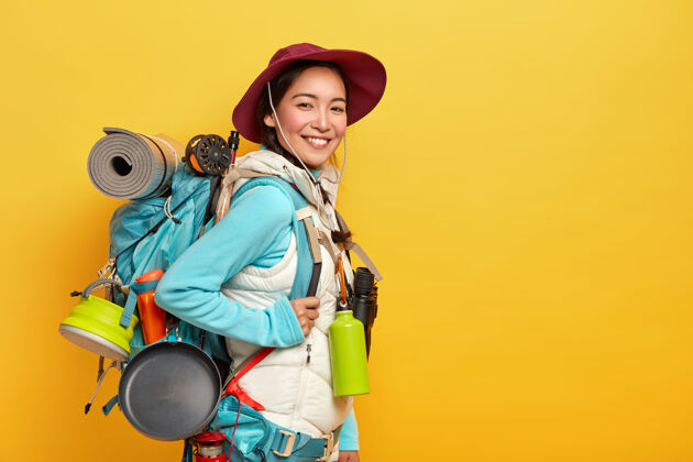 徒步旅行快乐的深色头发的亚洲旅行者背着大大的旅游背包 用望远镜旅行 靠着黄色的墙 戴着时髦的帽子 穿着背心的套头衫帽子看方向