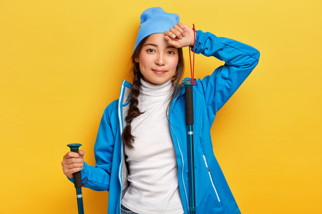 小径疲惫的亚洲女远足者拿着登山杖摆姿势 参加户外活动 旅行 穿着蓝色套装 摸摸额头 表情平静 隔着黄色的墙壁 与世隔绝休闲高领毛衣活动