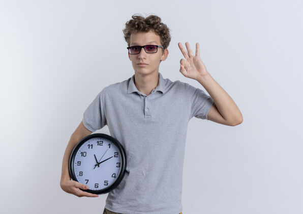 时钟一个穿着灰色马球衫的年轻人拿着墙上的挂钟 在白色上面显示ok标志标志马球墙壁