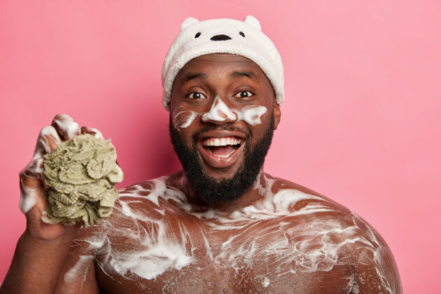 洗漱滑稽的黑胡子男人洗他的躯干 身体和脸上有泡沫 开心地笑 拿着海绵 戴着浴帽 隔离在粉红色的背景上皮肤海绵早晨