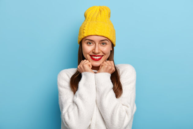 温暖好看的女人的肖像看起来是正面的 愉快的微笑 穿着红色的口红 有黑色的头发 黄色的帽子 白色的毛衣快乐笑声反应