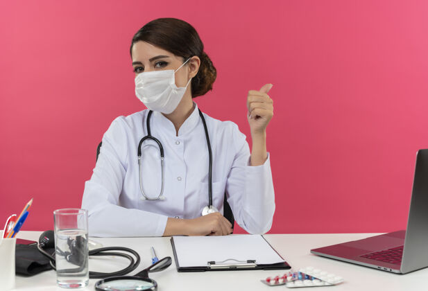专业自信的年轻女医生穿着医用长袍 戴着听诊器和口罩 坐在办公桌旁 手持医疗工具和笔记本电脑 竖起大拇指年轻拇指冠状病毒