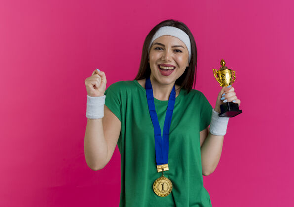 周围快乐的年轻运动女性戴着头带和腕带 脖子上戴着奖牌 手里拿着冠军杯 看起来在做“是”的手势女子穿年轻