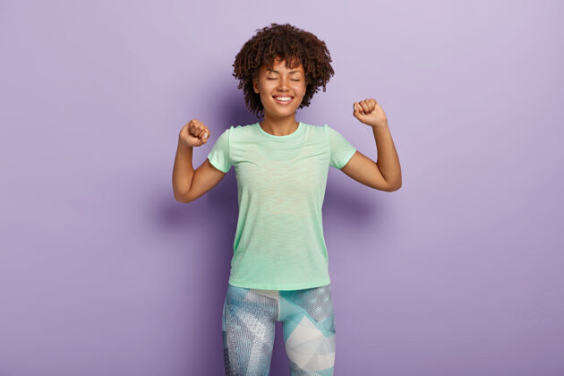 室内积极的年轻卷发女孩伸展手臂 感到高兴 做运动 穿休闲t恤和紧身裤强壮运动女性