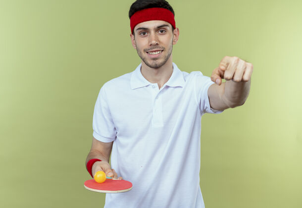 指着戴着头巾 拿着球拍和乒乓球的年轻人微笑着用食指指着你站在绿色的墙上桌子球拍球