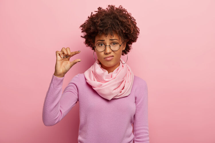 不满意心烦意乱的卷发女人塑造了一个小小的手势 展示了一些非常微小的东西 戴着眼镜 休闲套头衫和围巾小人眼镜